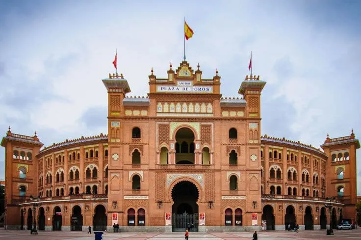 The Plaza de Toros de las Ventas bullring with its grand facade, arched entrances, and Spanish flag flying at the top under a cloudy sky.