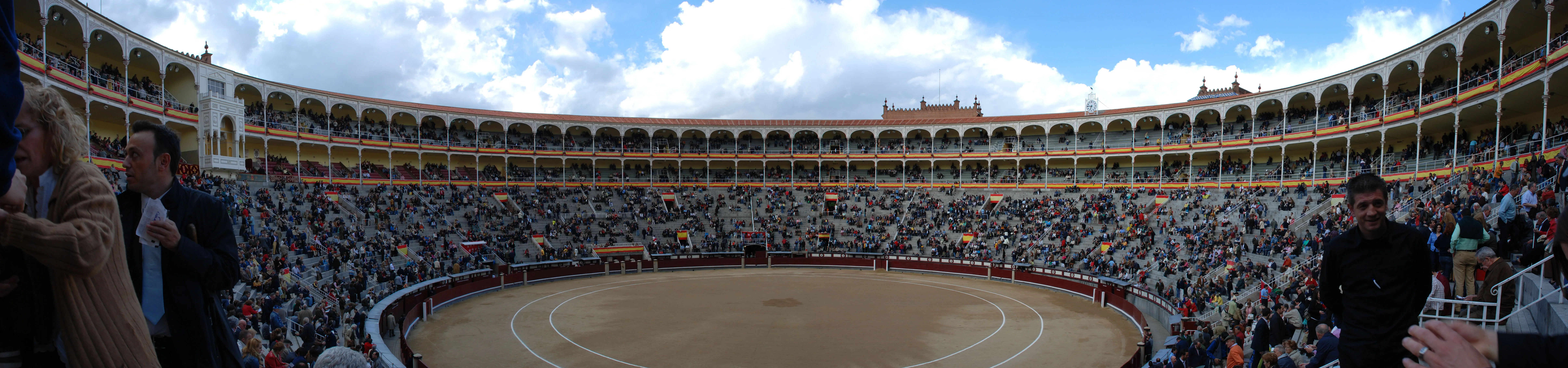 Panoramic view of the Plaza de Toros de las Ventas bullring with spectators in the stands and a sandy arena under a partly cloudy sky