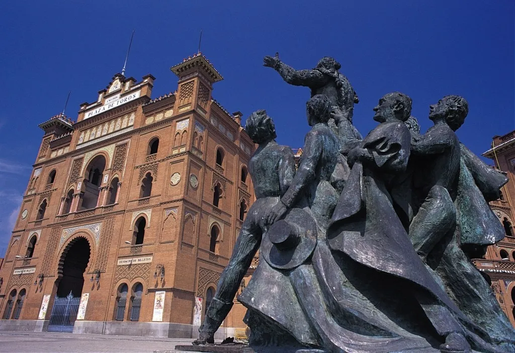 Bronze statue of bullfighters in front of the brick facade of the Plaza de Toros de las Ventas under a blue sky