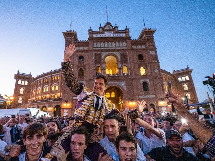 A matador in ornate costume is lifted by a crowd in front of the historic Plaza de Toros de las Ventas building at dusk.