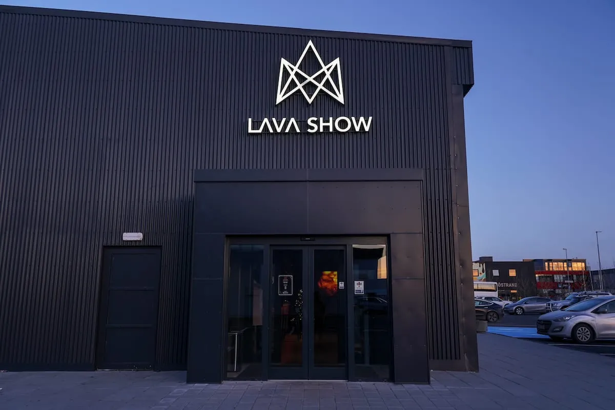 Fasada budynku Lava Show wykonana z ciemnej, falistej blachy, z podświetlanym białym geometrycznym logo i napisem 'LAVA SHOW' nad wejściem; po prawej stronie widoczny parking o zmierzchu.