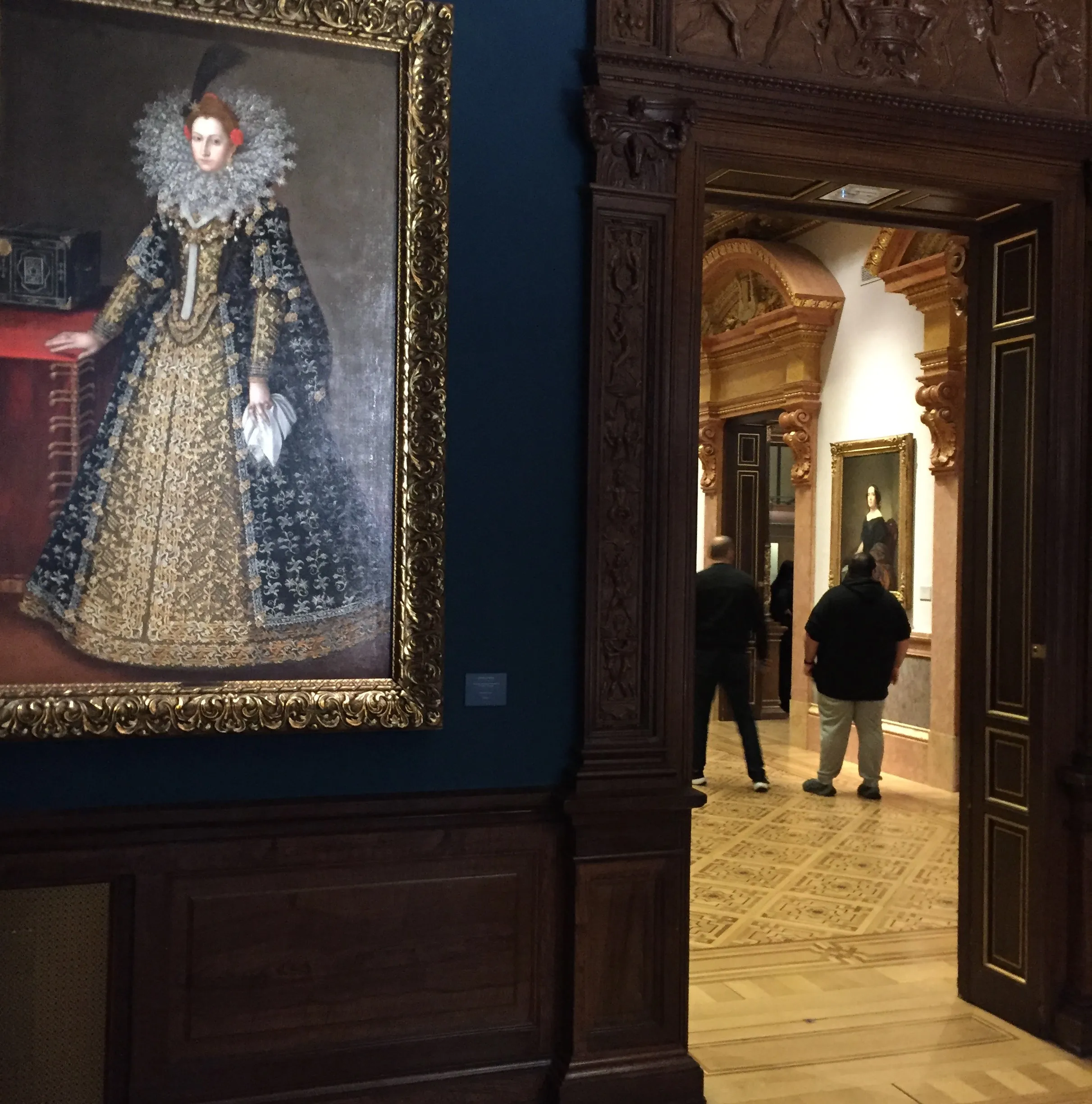 Un gran retrato pintado de una mujer con atuendo histórico ornamentado cuelga en una pared azul oscuro en una galería, mientras los visitantes atraviesan una puerta de madera hacia una sala contigua.