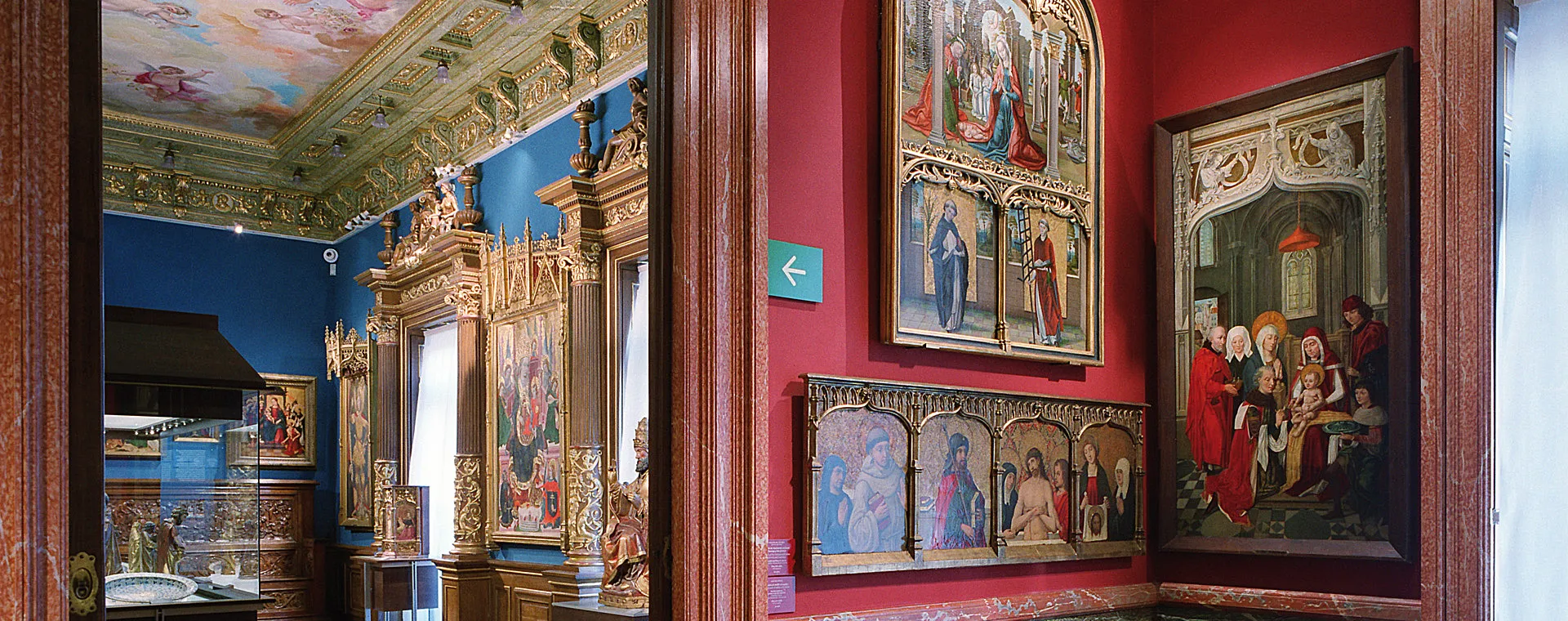 Galería interior con paredes rojas, retablos de madera ornamentados y pinturas religiosas en marcos dorados