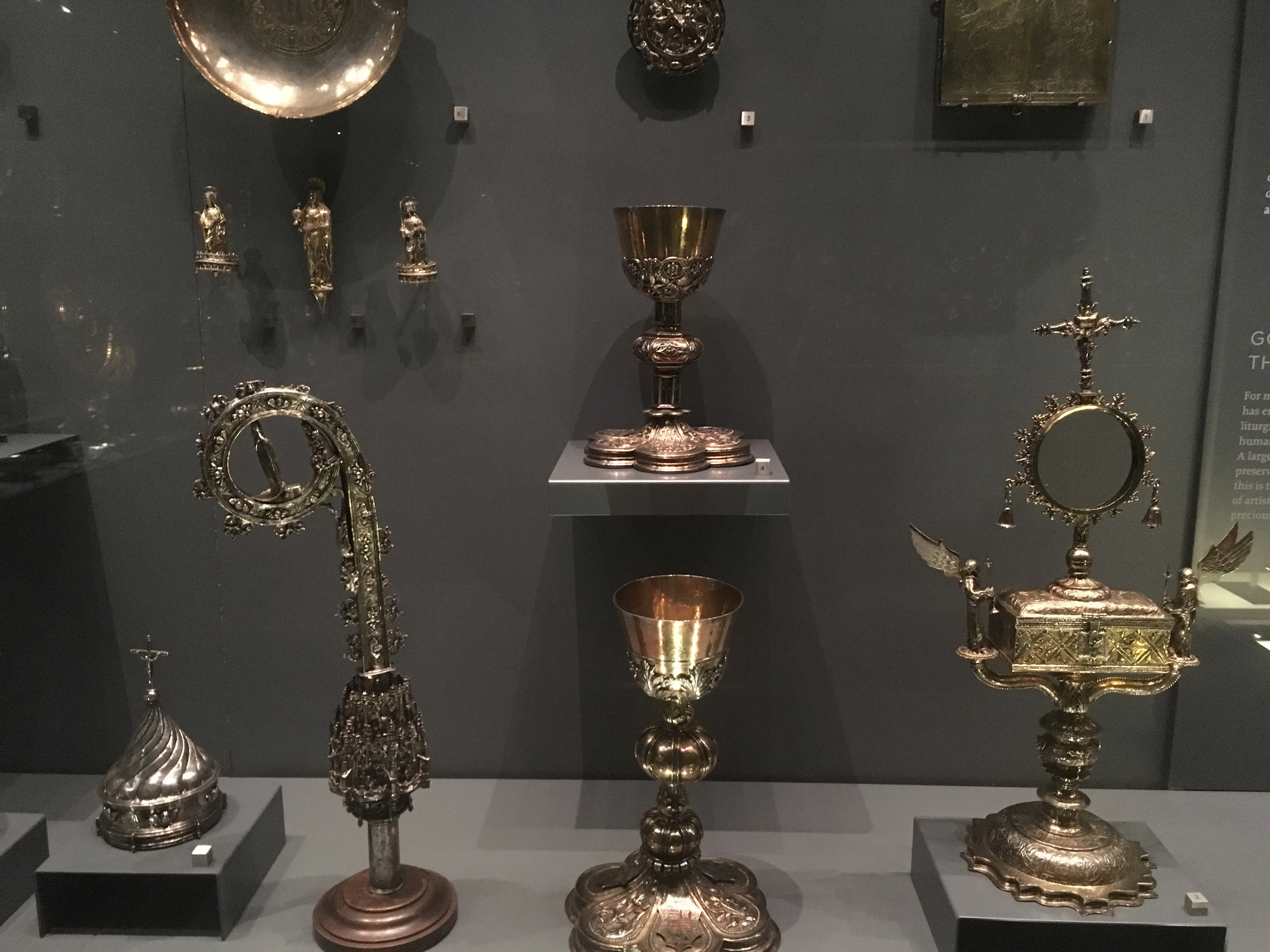 Artefactos religiosos dorados que incluyen cálices, un ciborio y bastones ornamentados, exhibidos sobre pedestales en una galería de museo