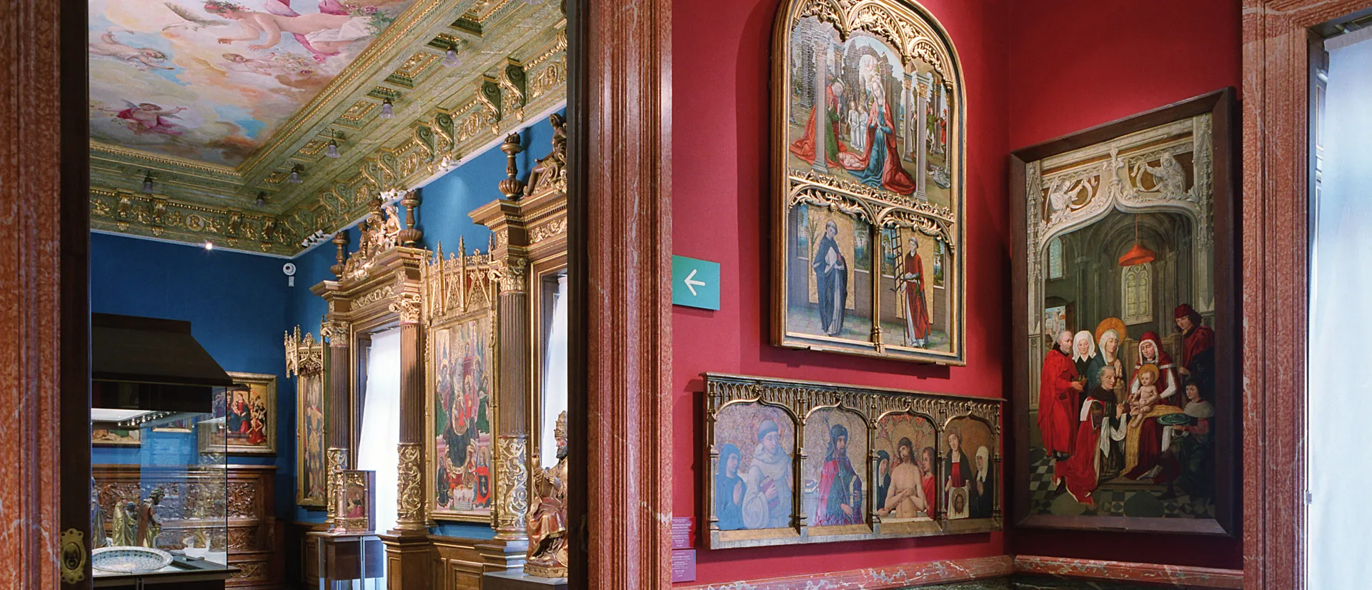Galería interior con paredes rojas que exhiben pinturas religiosas y una pared azul con un retablo dorado