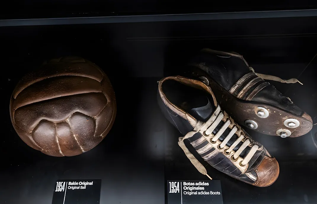 A vintage brown football and a pair of old black football boots with white laces on display in a dark case.
