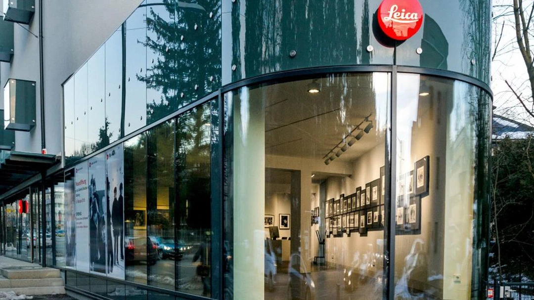 Buitenaanzicht van Leica Gallery Salzburg met een gebogen glazen gevel, een rood, rond Leica-logo en ingelijste foto's die zichtbaar zijn in de galerieruimte.