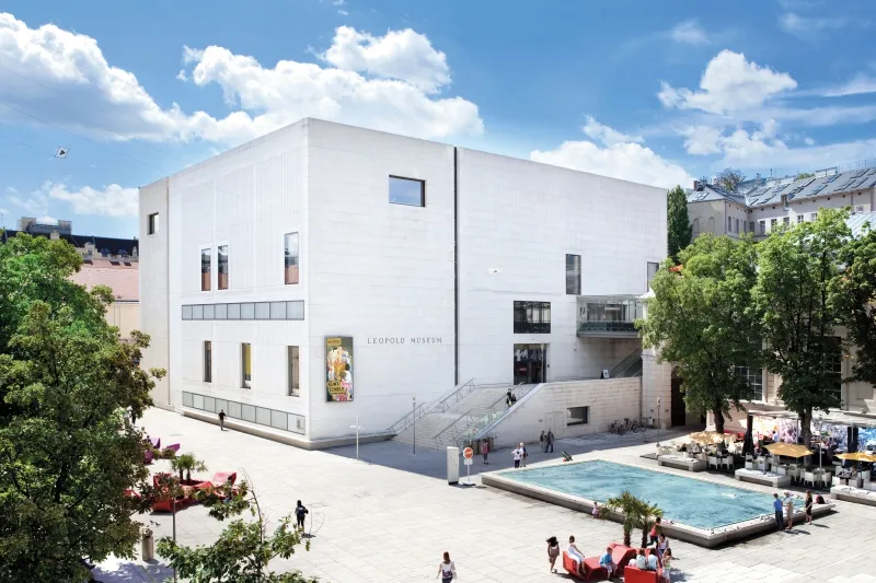 Wit modern gebouw met grote ramen en een bord met 'Leopold Museum' erop, onder een blauwe hemel met wolken
