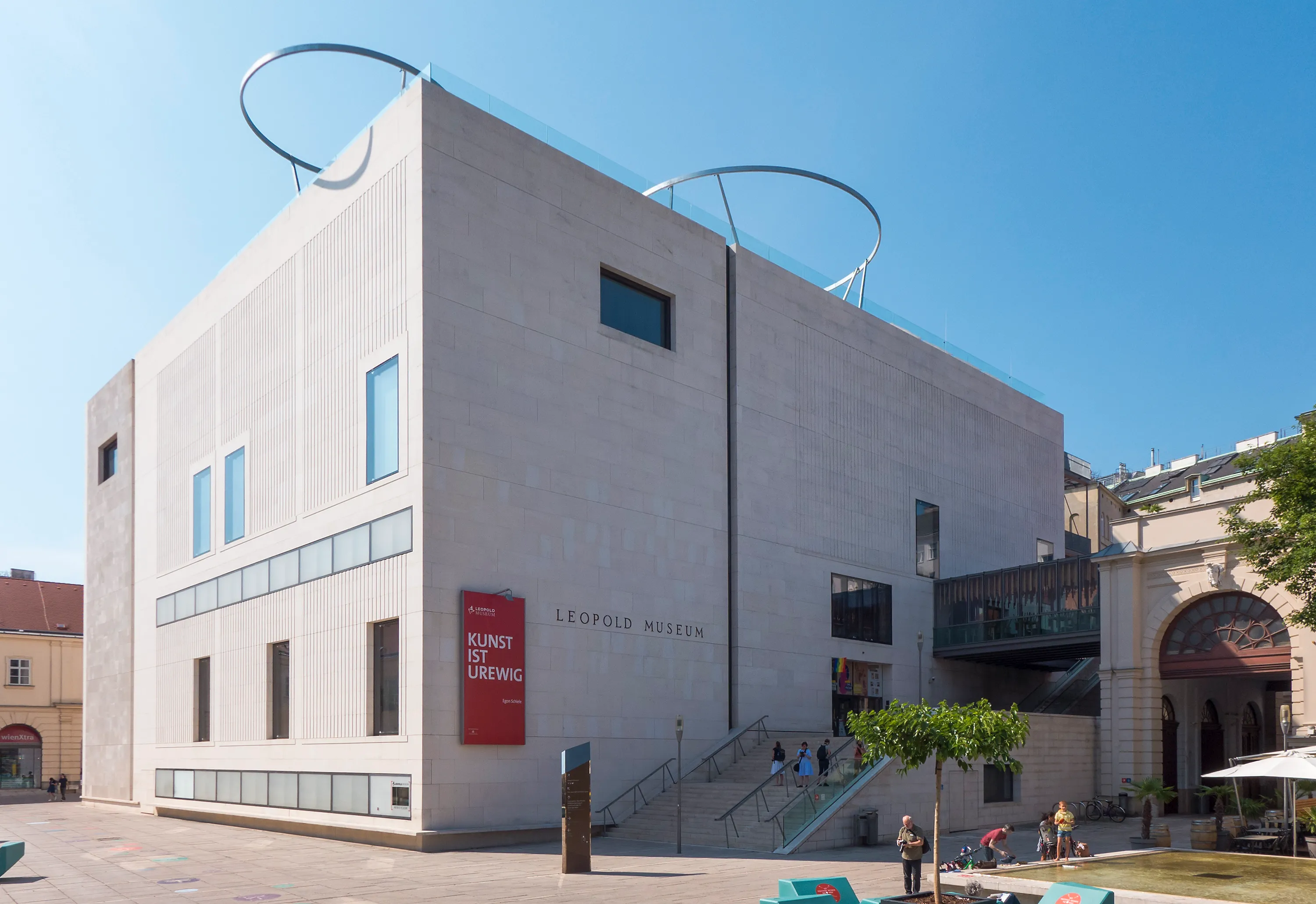 Het Leopold Museum van buitenaf, met een moderne lichtgekleurde gevel, cirkelvormige metalen sculpturen op het dak en een rood bord met de tekst 'KUNST DER LIEBE'
