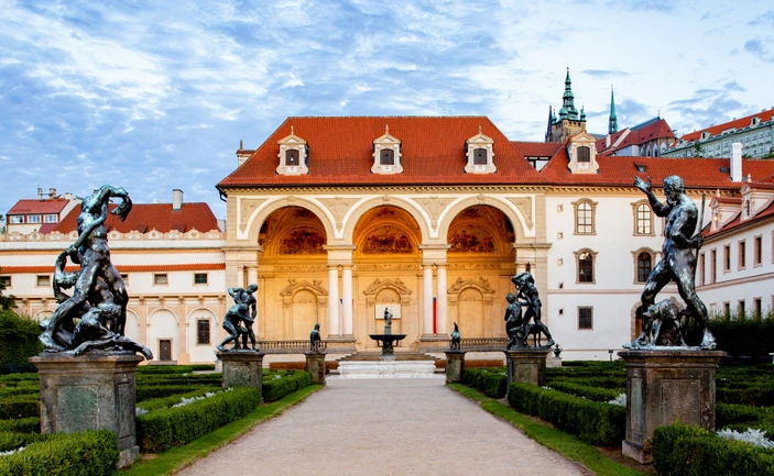 Un edificio majestuoso con techo rojo y fachada arqueada, flanqueado por estatuas en un jardín formal.