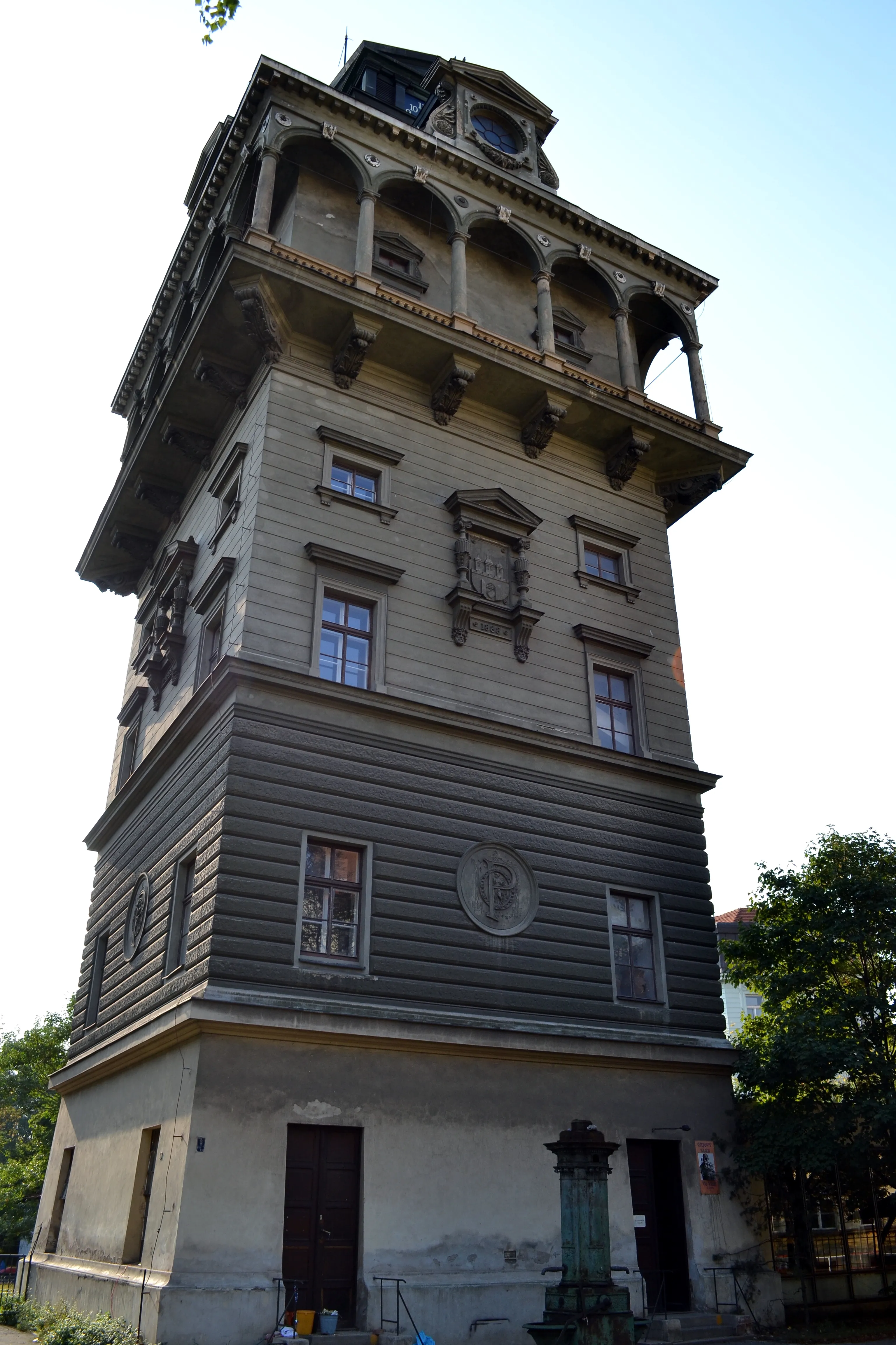 Una torre de agua histórica y alta con intrincados detalles arquitectónicos y una esfera de reloj, rodeada de vegetación bajo un cielo despejado