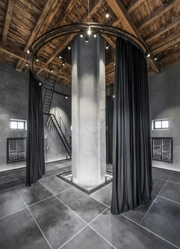 Interior de la Torre de Agua de Letná con una columna central de hormigón, cortinas negras, vigas de madera expuestas y una escalera de caracol.