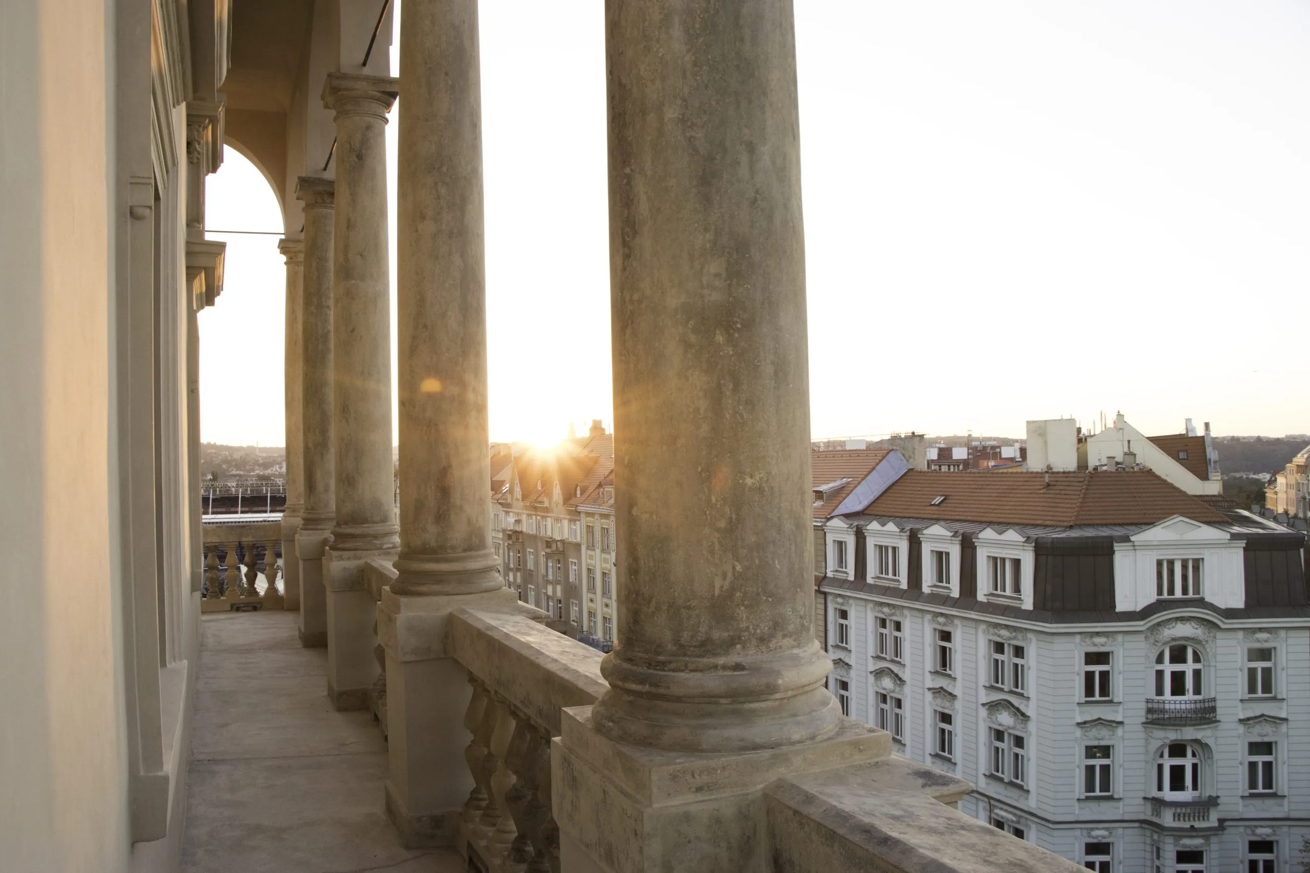 columnas de piedra de un balcón con vistas a una ciudad con edificios y un sol poniente