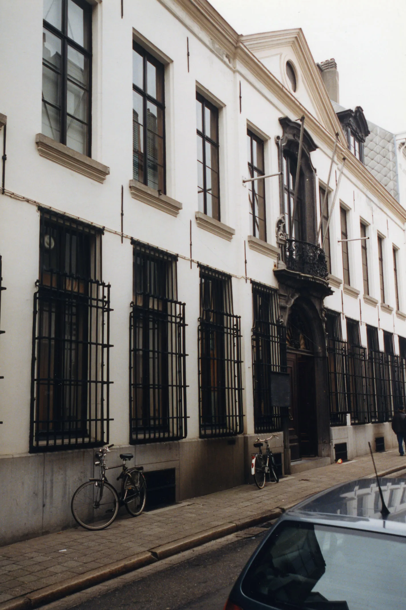 Fachada de un edificio blanco de varias plantas con rejas de hierro negro en las ventanas, un pequeño balcón sobre una entrada arqueada y bicicletas aparcadas en la acera