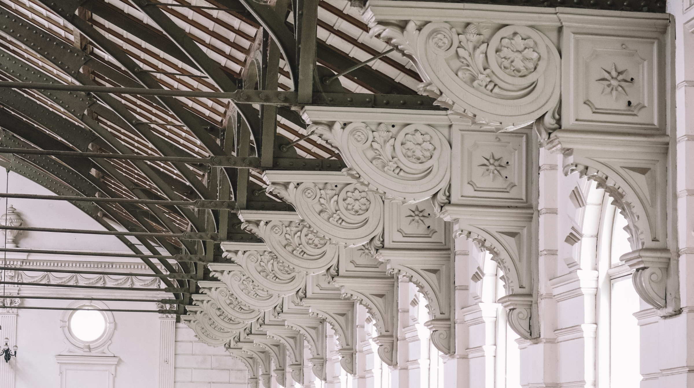 Ornate witte consoles met bloemmotieven die een gebogen metalen en glazen dakstructuur ondersteunen in een historisch gebouw