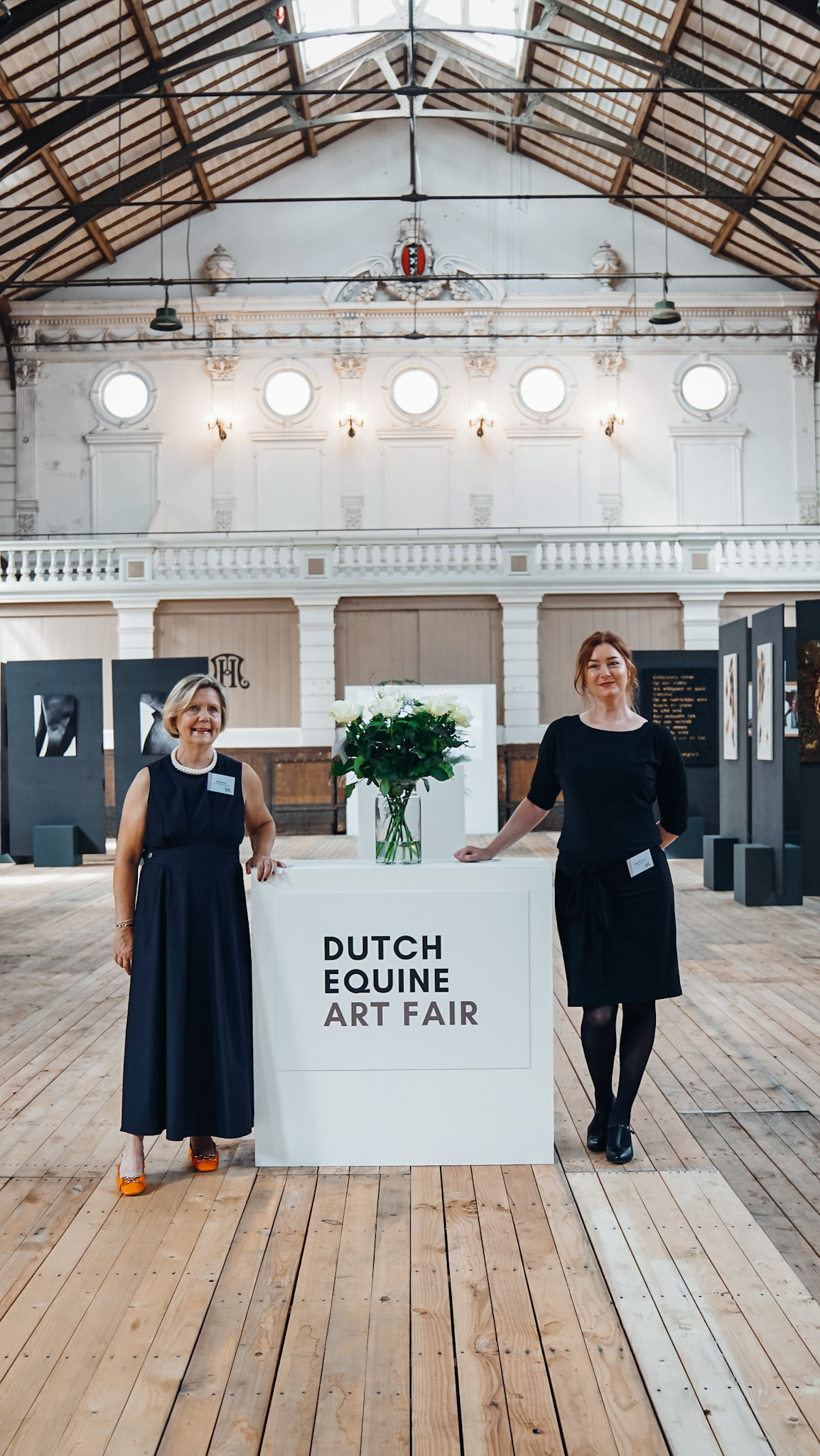 Dwie kobiety stoją obok białego lady, na którym widnieje napis 'DUTCH EQUINE ART FAIR', w przestronnej sali wystawowej z wysokimi sufitami i drewnianą podłogą