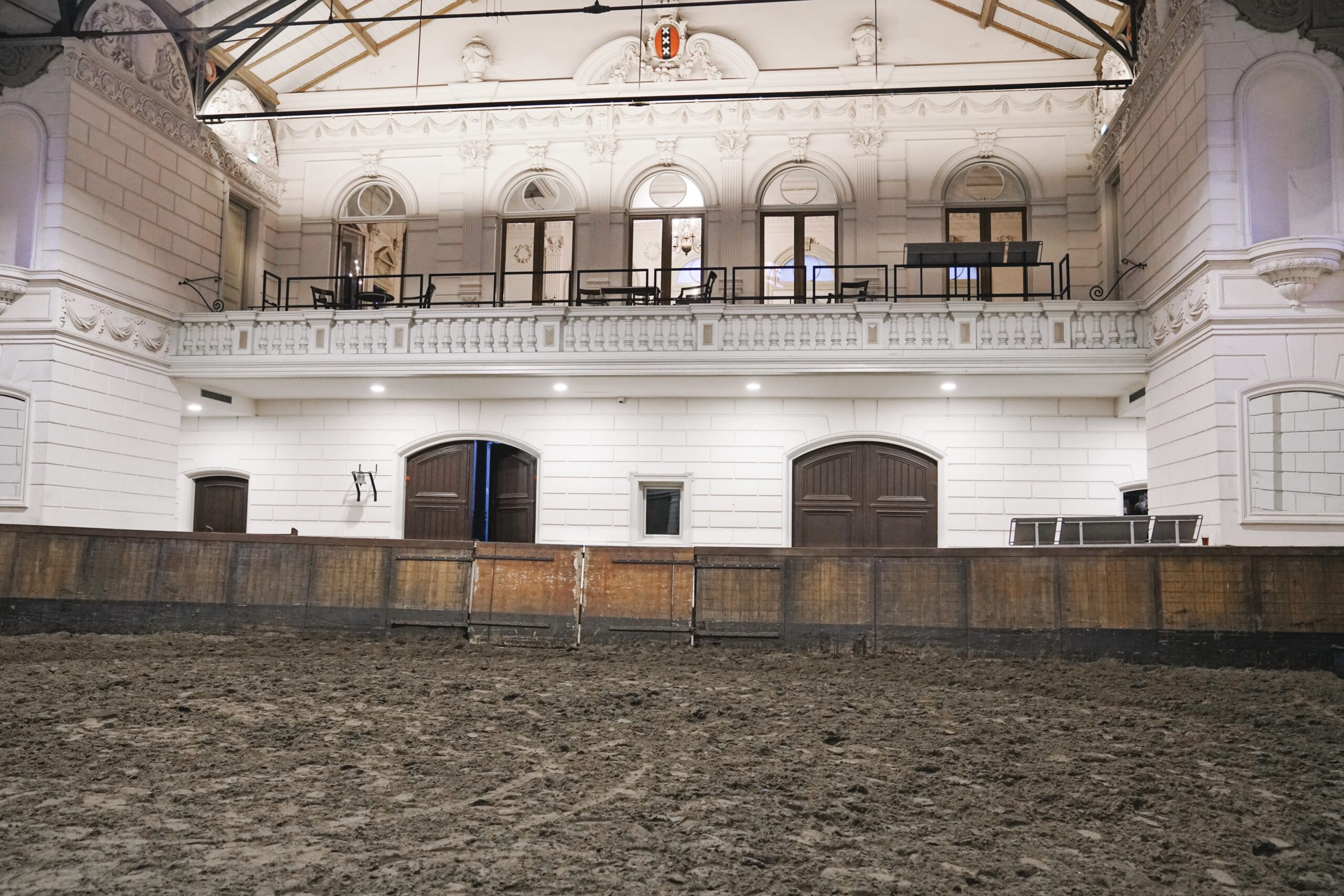 Arena halowa z glinianą podłogą i drewnianymi barierami, białe ściany z łukowymi oknami i balkonem powyżej, ozdobne belki stropowe