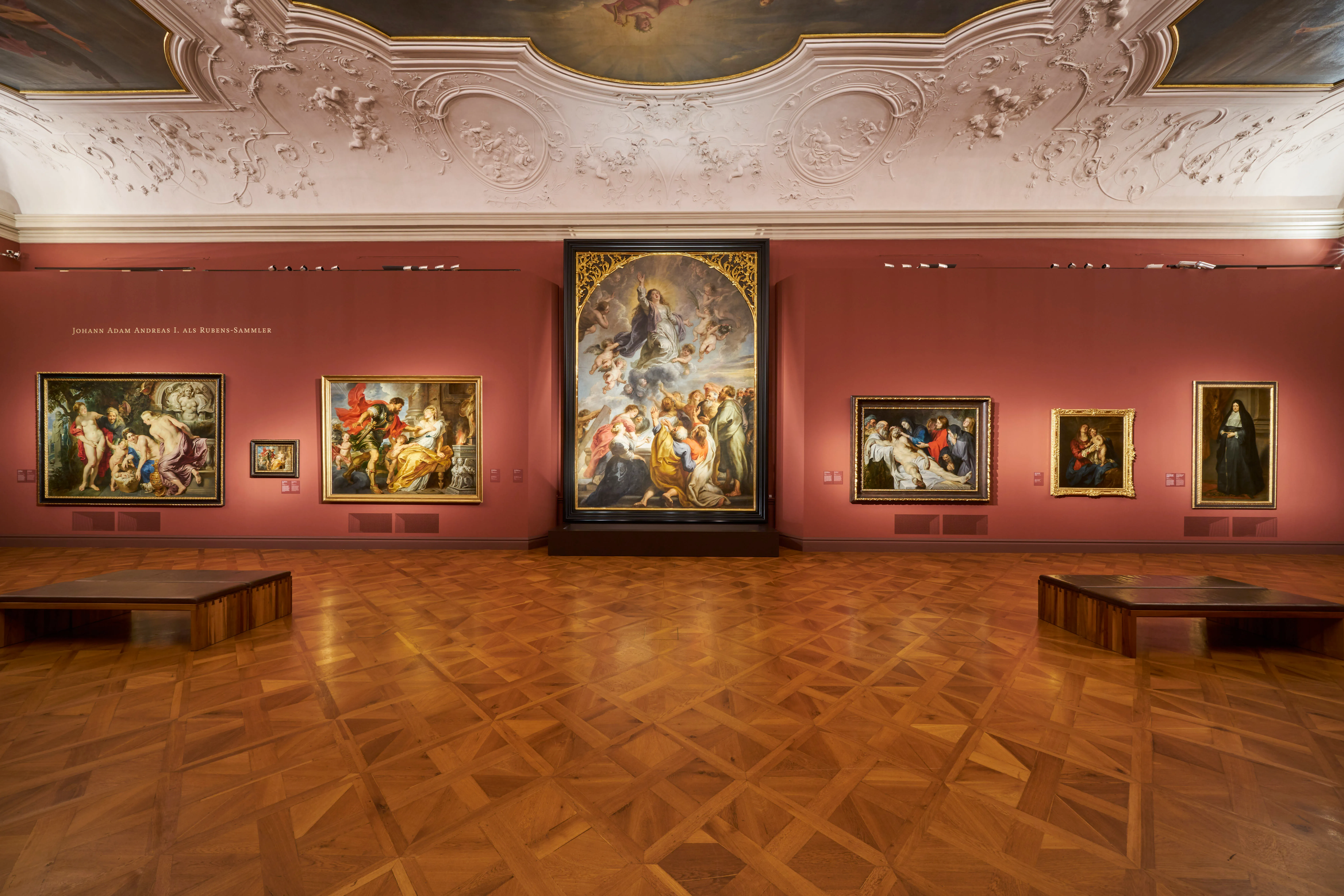 Una sala de galería con techo ornamentado, paredes rojas y múltiples pinturas en exhibición