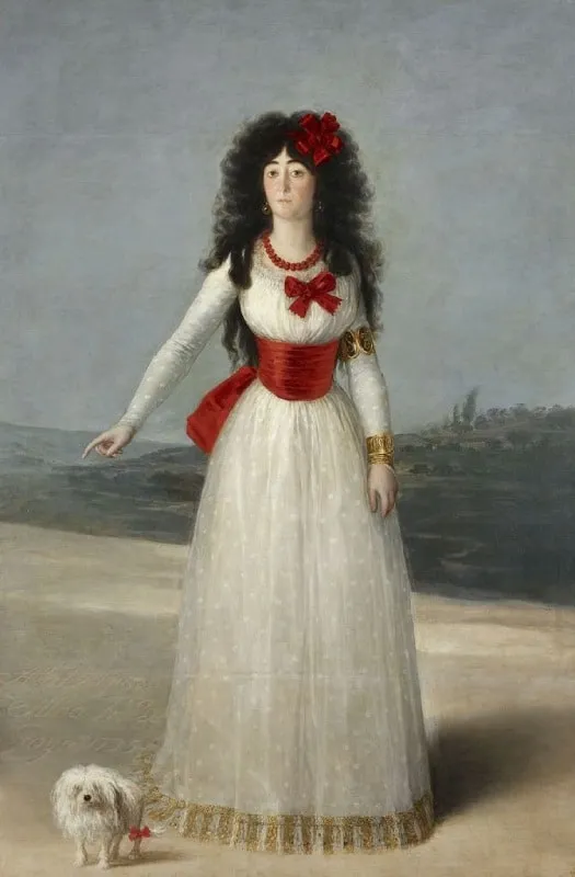 Una mujer con un vestido blanco con detalles rojos y una flor roja en el pelo, de pie con un perro blanco pequeño