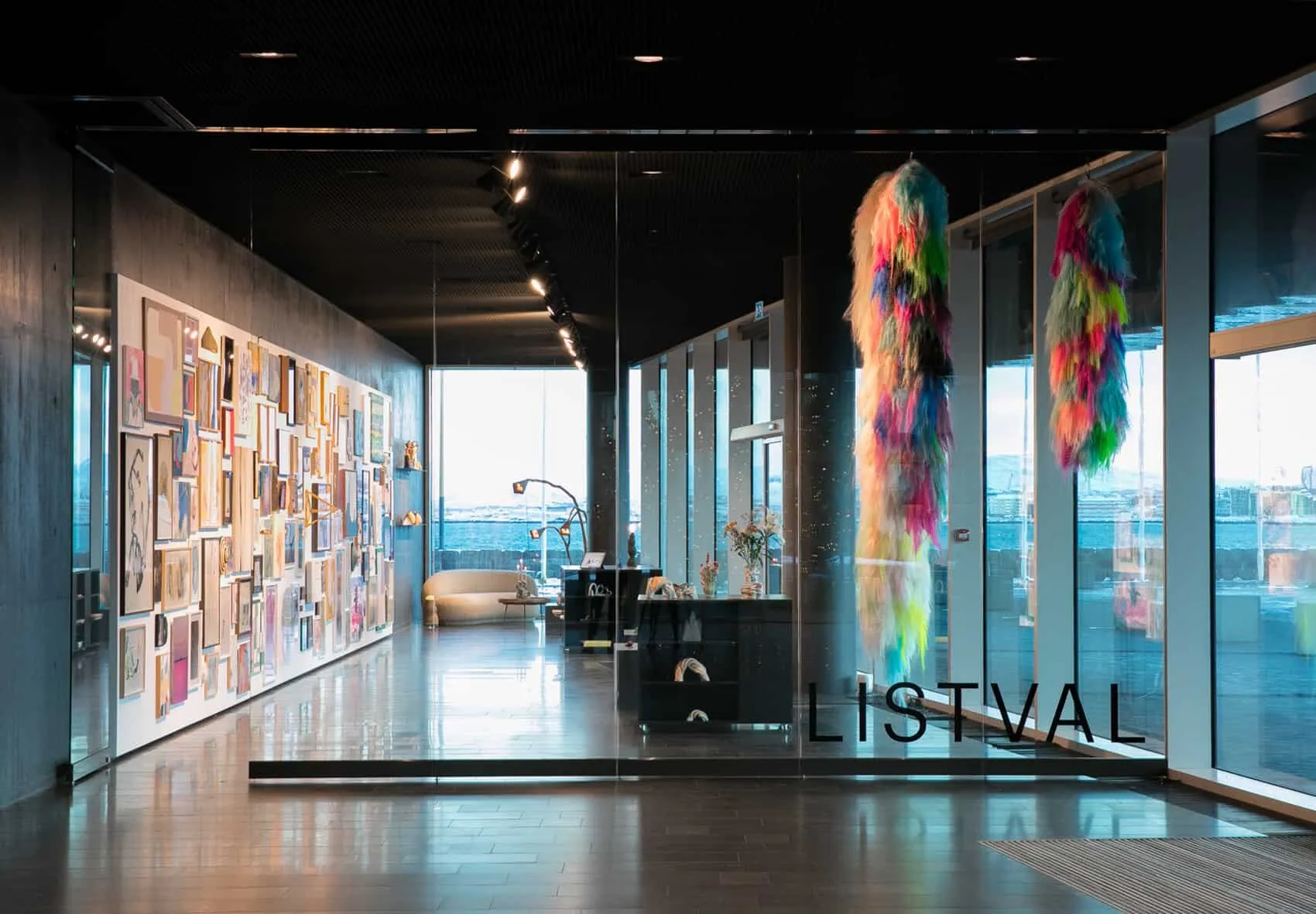 Wnętrze Listval Gallery z ścianą wypełnioną oprawionymi dziełami sztuki, kolorowymi wiszącymi instalacjami z tkaniny przy dużych oknach oraz nazwą galerii widoczną na szklanej ścianie działowej.