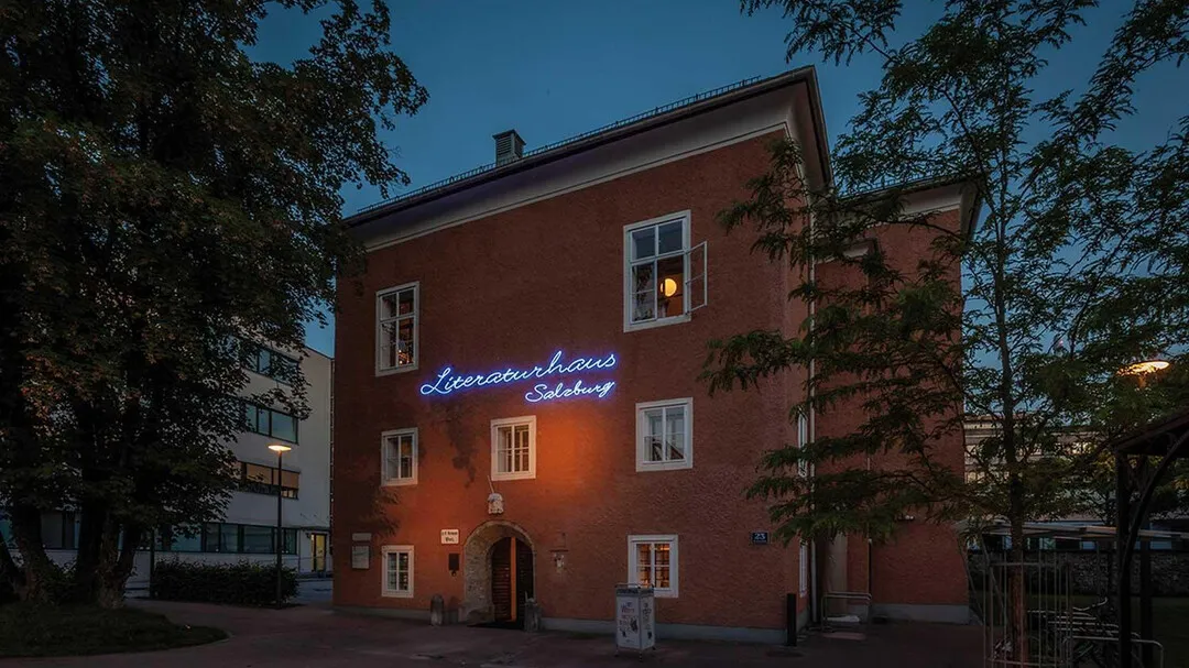 Edificio de ladrillo rojo al atardecer con un letrero de neón azul brillante que dice 'Literaturhaus Salzburg' en la fachada, enmarcado por árboles y con luz cálida en las ventanas.