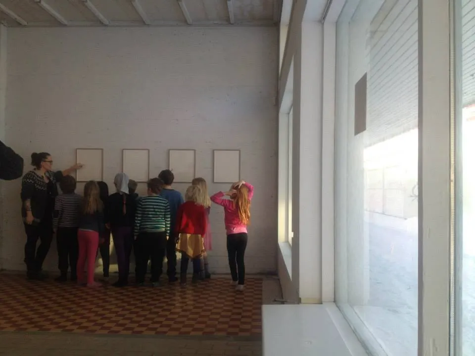 Een groep volwassenen en kinderen staat in een galerieruimte en kijkt naar vier wit ingelijste kunstwerken aan een muur.