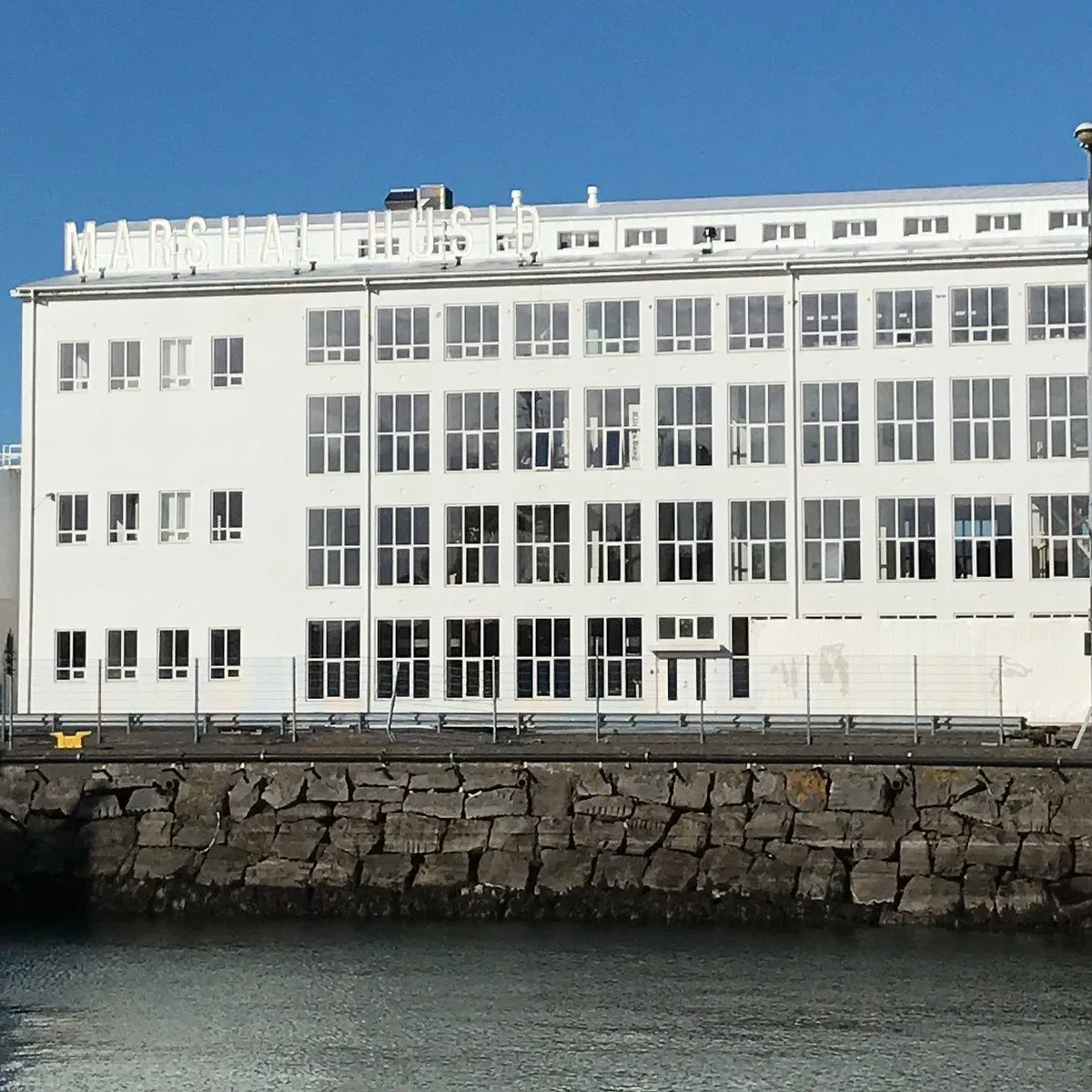 Wit, meerverdiepingsgebouw Marshall House met grote vensters met roedeverdeling langs een rotsachtige waterkant onder een helderblauwe lucht