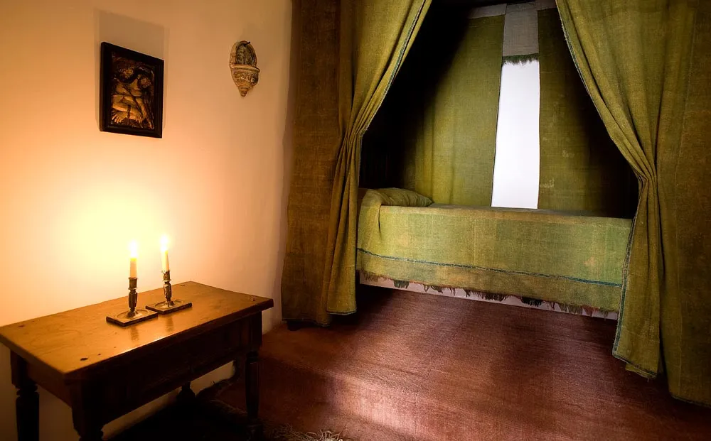 A wooden table with two candlesticks, a framed picture on the wall, and a bed with green drapery in a dimly lit room
