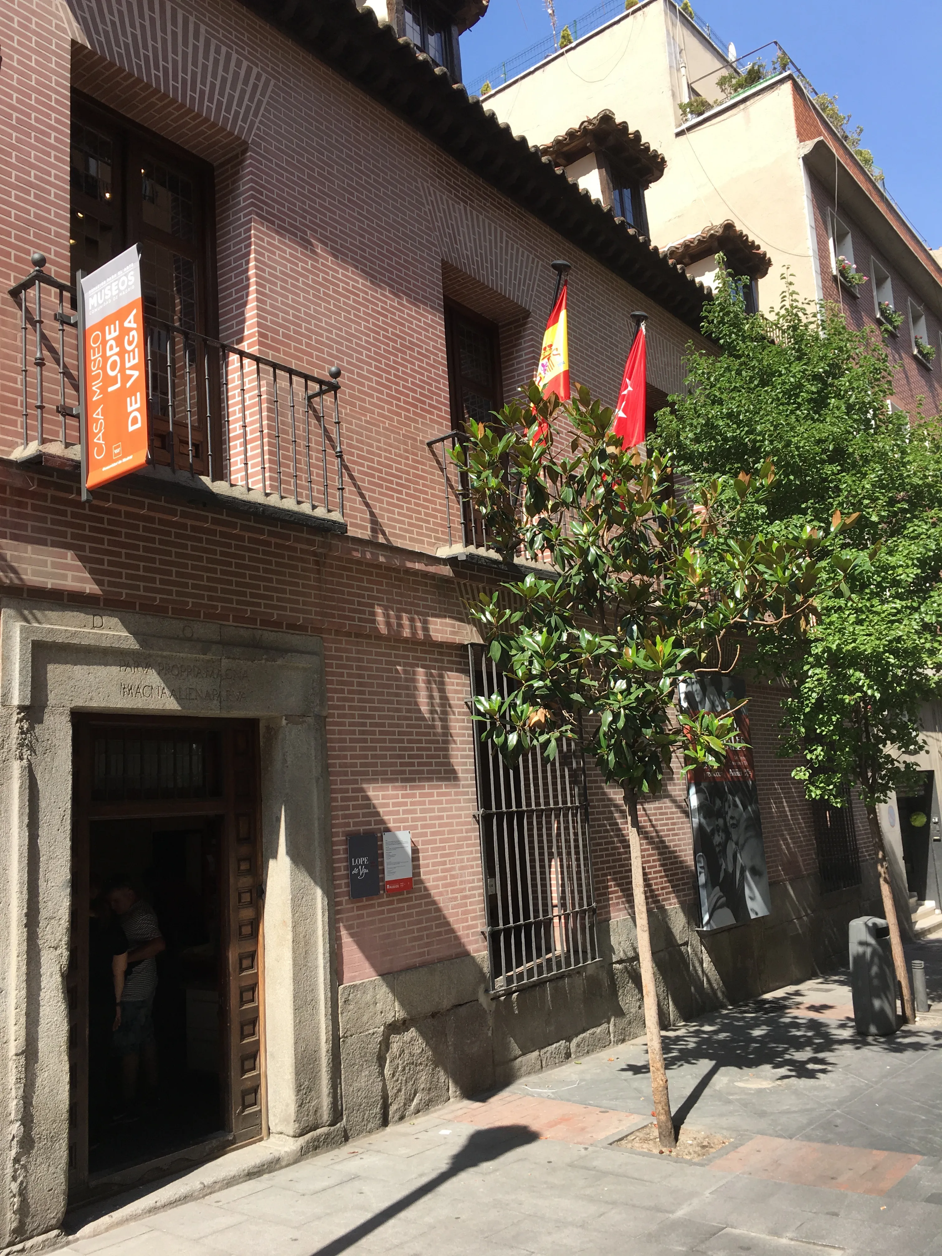 Brick building with a stone entrance and a sign reading 'Casa Museo Lope de Vega' on the facade, with Spanish and red flags displayed, and a tree on the sidewalk