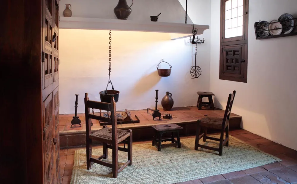 Interior of a historic kitchen with a central hearth, wooden chairs, and traditional cooking implements