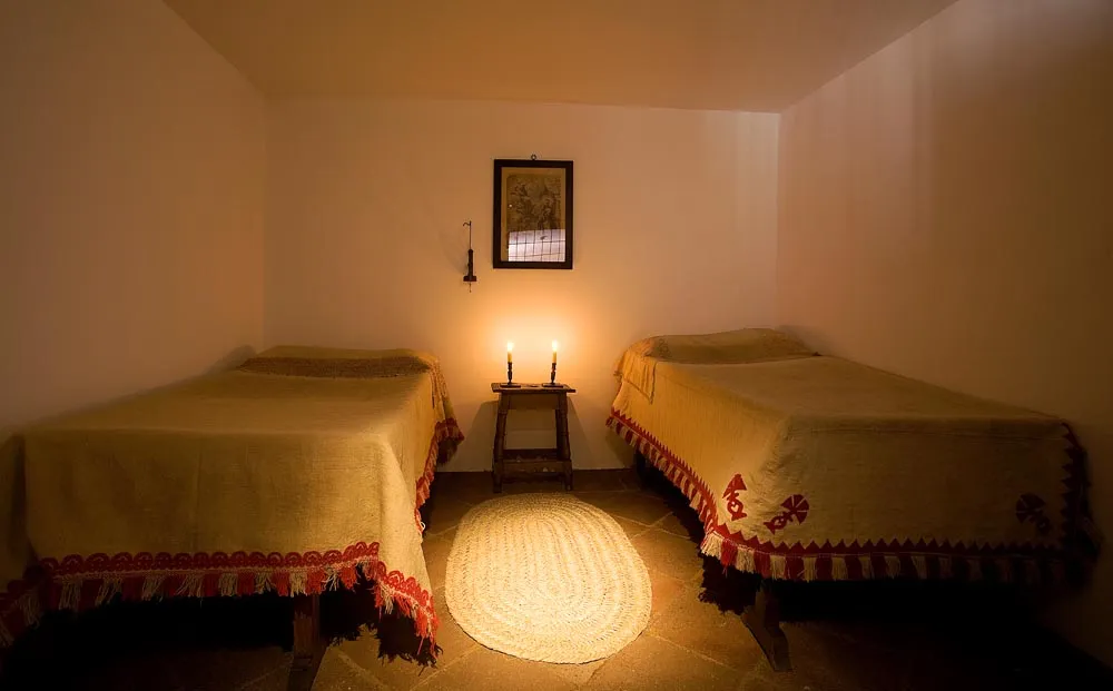 A dimly lit room with two beds covered in yellow linens and red trim, a small table with candles, and a framed picture on the wall.