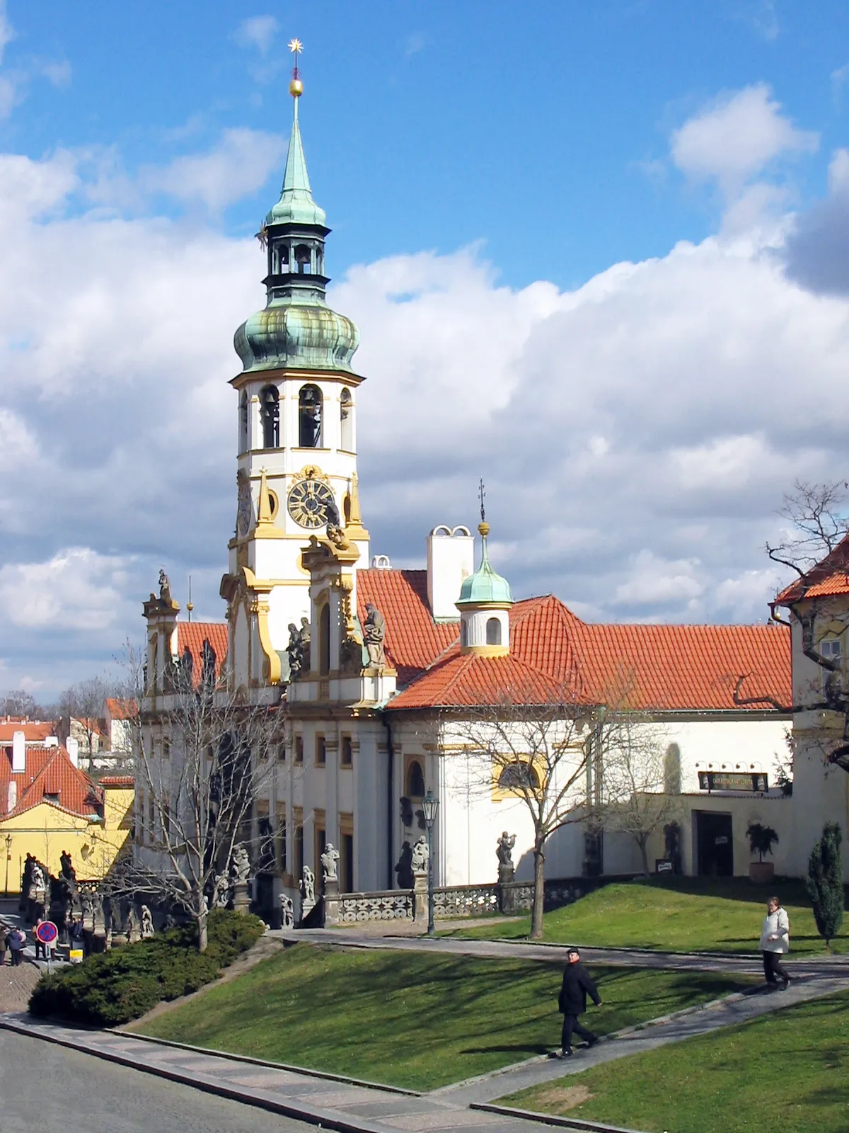 Barokowa wieża z zieloną iglicą i zegarem, biały budynek z czerwonym dachem i zielonym trawnikiem z przodu