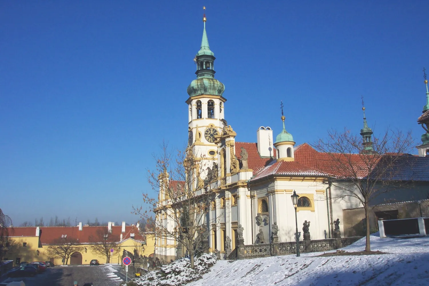 Barokke kerk met een groene spits en kerktoren in Loreta in Praag, met sneeuw op de grond en een helderblauwe lucht