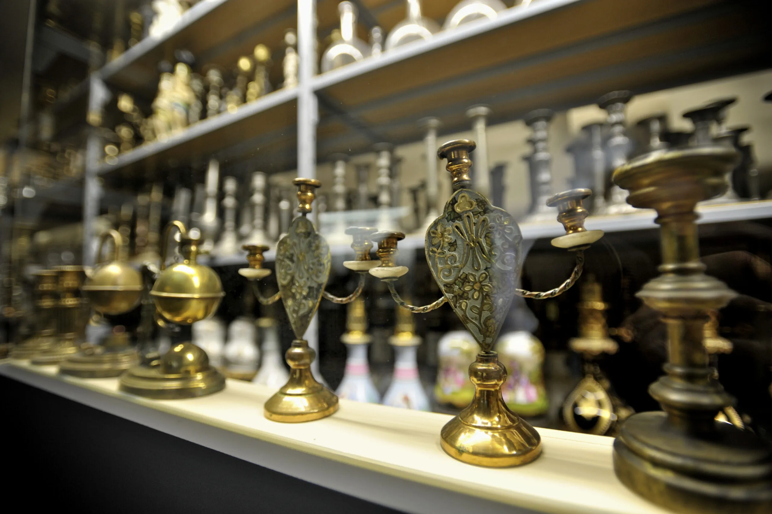 Candelabros de latón ornamentados y lámparas históricas dispuestos en estantes blancos en una vitrina de museo, con filas de objetos de iluminación adicionales visibles al fondo.