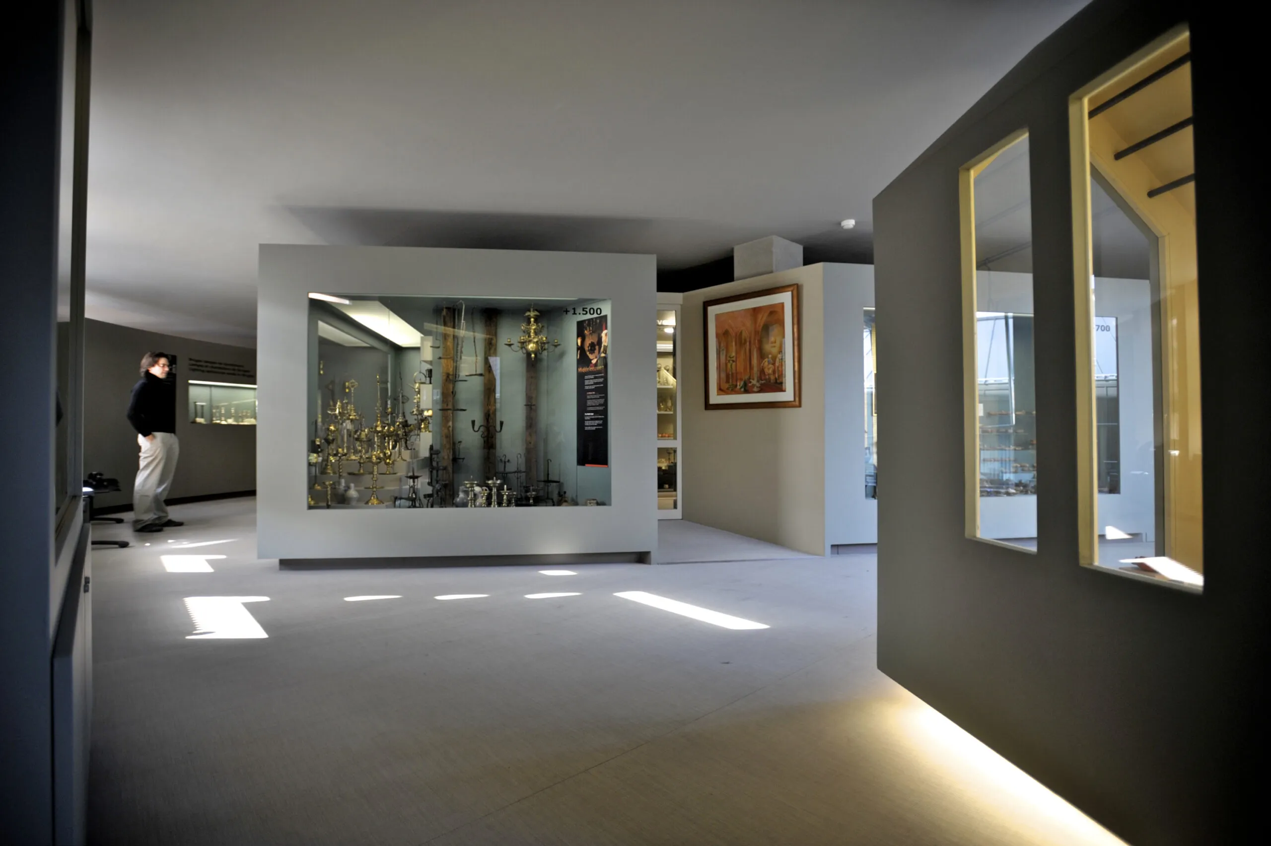 Interior de un museo con paredes grises, una gran vitrina de cristal que contiene lámparas de araña y accesorios de iluminación, obras de arte enmarcadas y un visitante observando la exposición