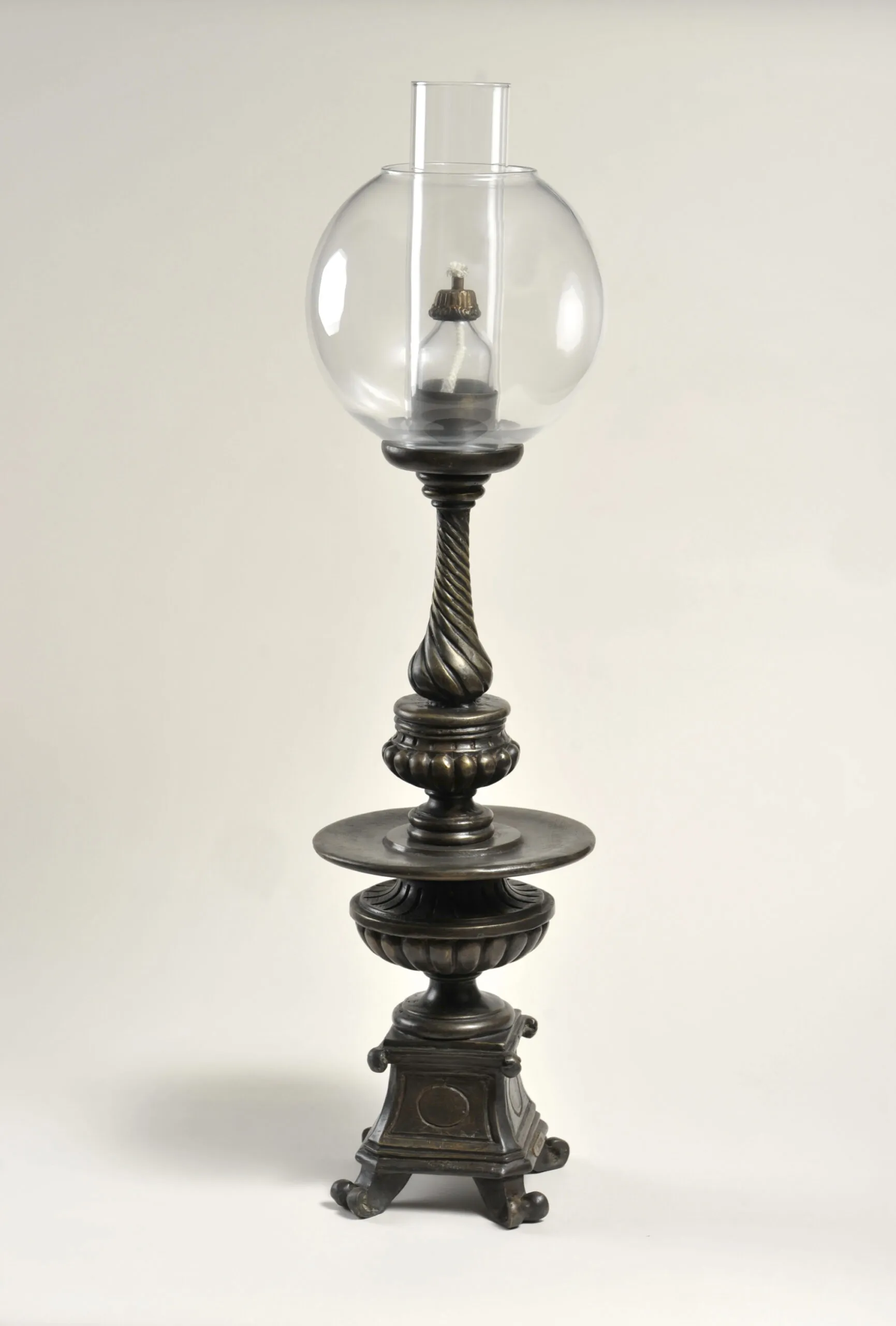 Lámpara de aceite antigua con globo de cristal transparente, vástago de metal oscuro trenzado y base de hierro fundido ornamentada con patas de voluta, fotografiada sobre un fondo blanco.