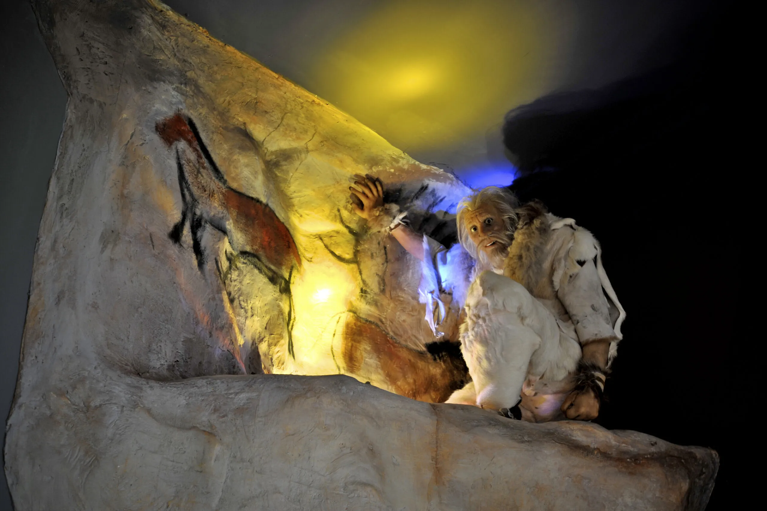 Diorama de museo que muestra a una figura prehistórica vestida con pieles de animal pintando animales en la pared de una cueva, iluminada dramáticamente con una cálida luz amarilla y azul.