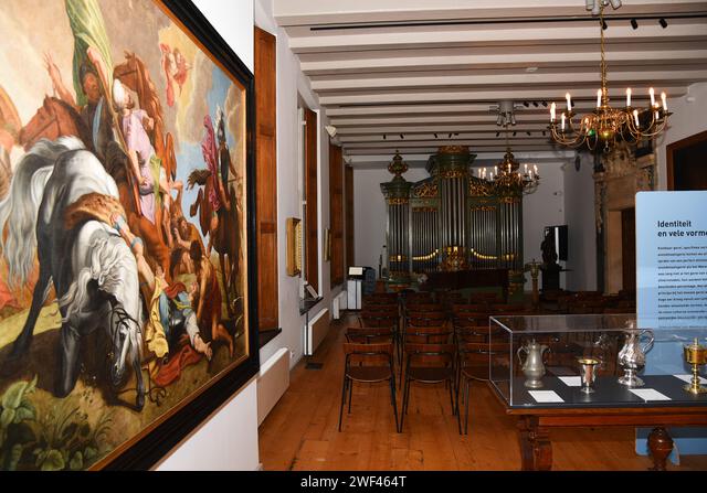 Long room with wooden floors and beams containing a large painting of figures on horseback, an organ, display cases, and seating arrangement