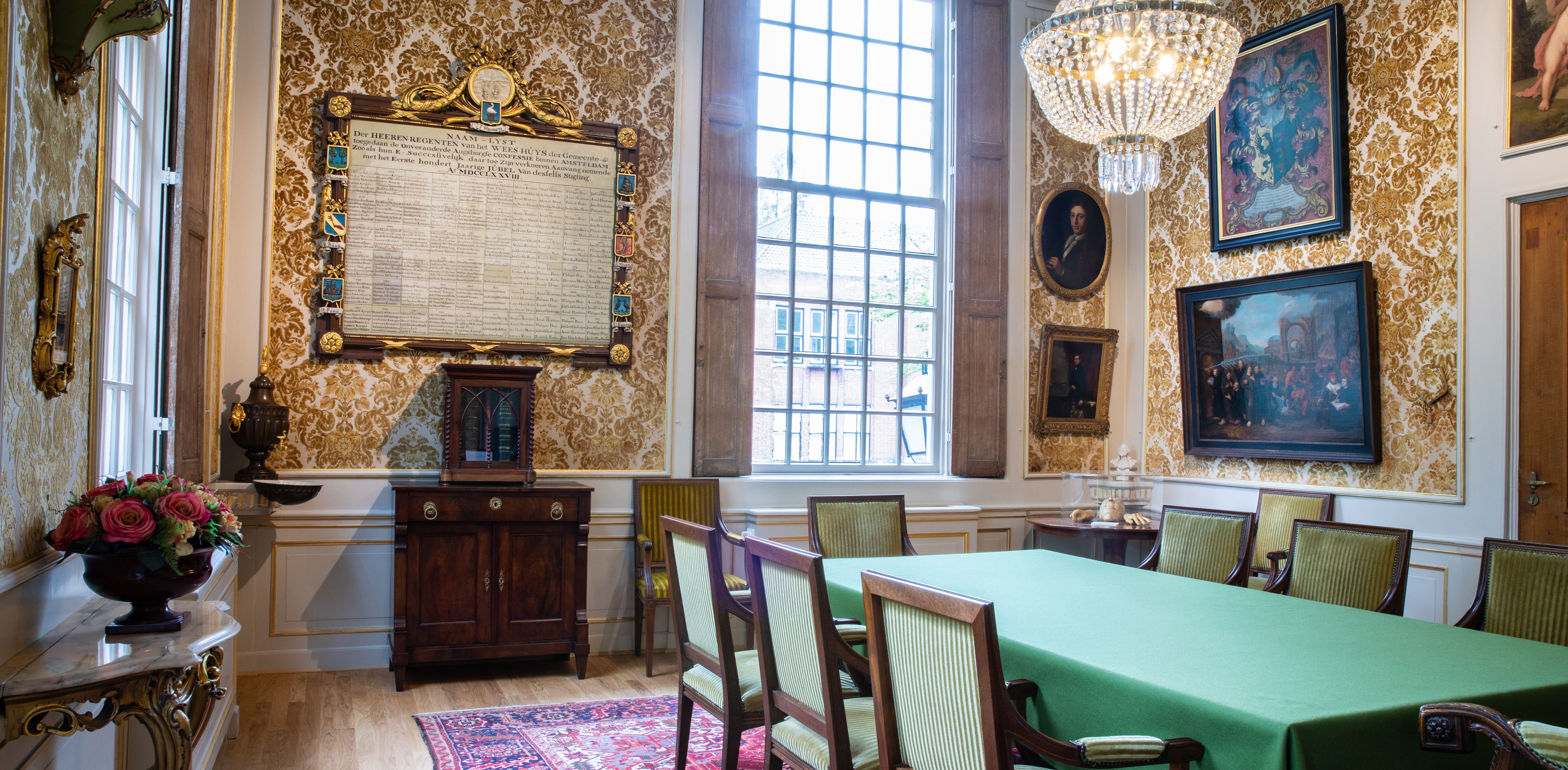 Elegante sala de reuniones con papel tapiz ornamentado en dorado, grandes ventanas con patrón de cuadrícula, mantel verde y obras de arte enmarcadas en las paredes