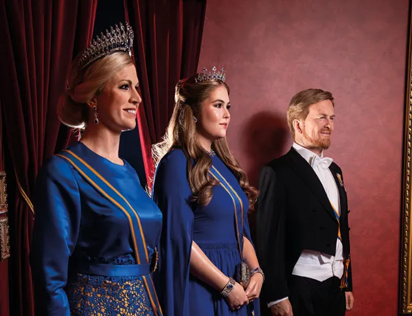 Drie wassen beelden van koninklijke figuren in formele kleding: twee vrouwen in blauwe jurken met tiara's en een man in een zwarte smoking voor een rode achtergrond.