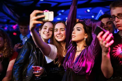 Drie vrouwen maken een selfie met een smartphone in een schemerige kamer met blauwe verlichting, omringd door andere bezoekers en wassenbeelden.