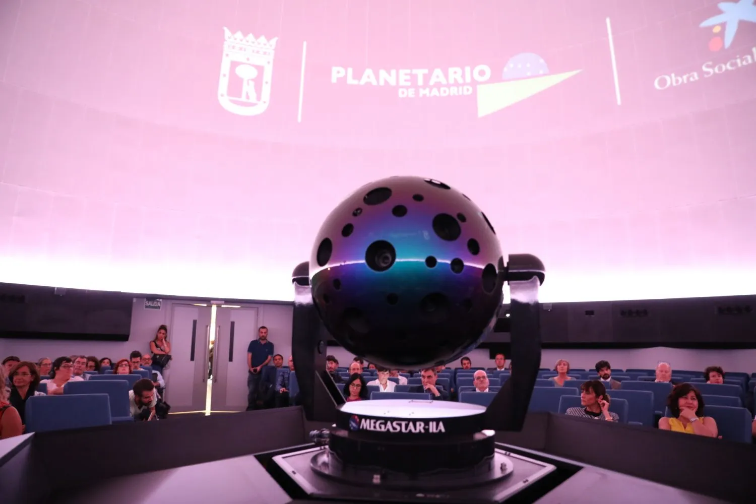 A large spherical projector labeled 'MEGASTAR IIA' in the center of a planetarium dome, with the 'PLANETARIO DE MADRID' logo projected on the curved ceiling and audience members seated in rows.