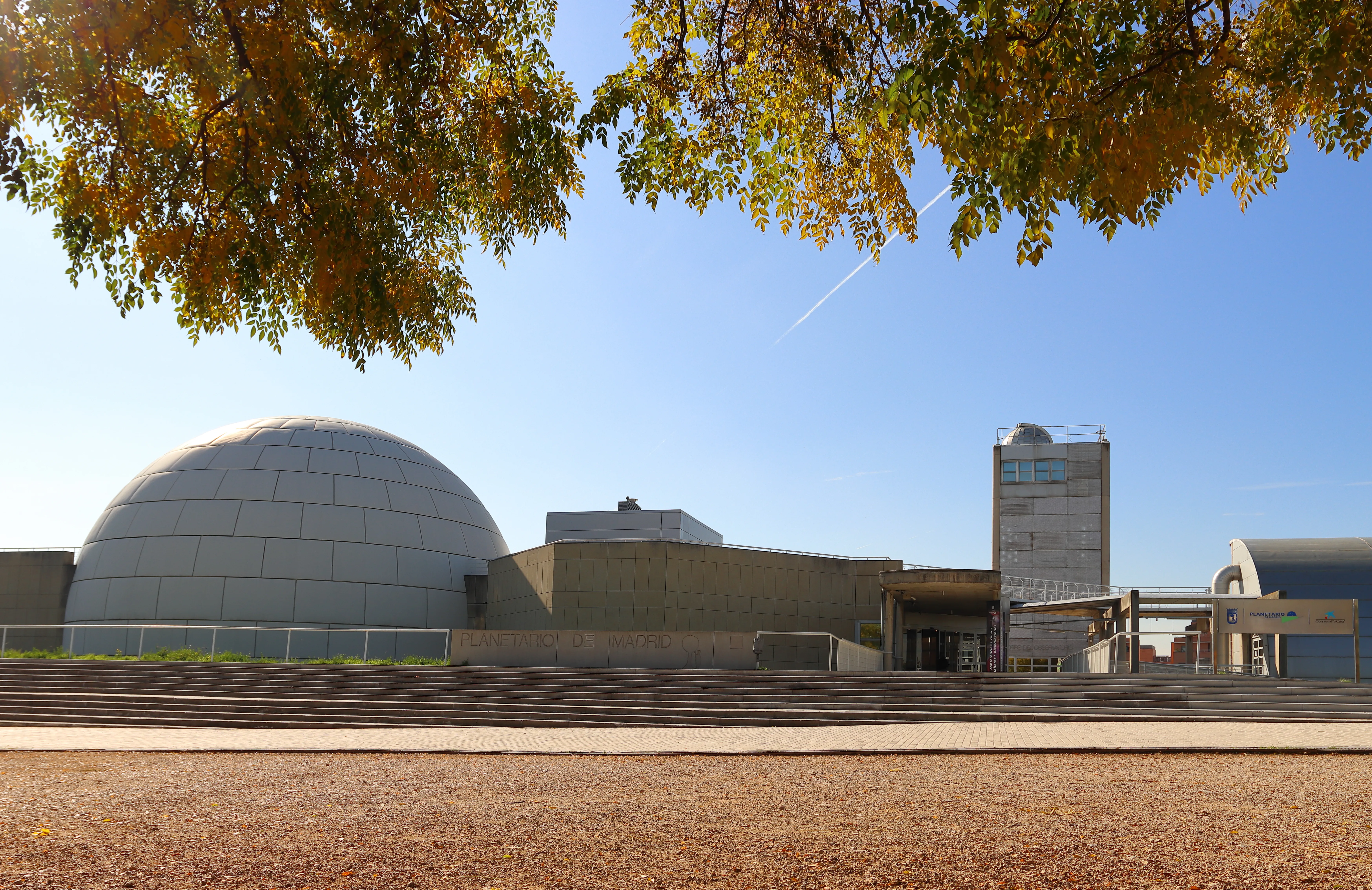 The Madrid Planetarium with a large dome and modern buildings under a blue sky with autumn trees in the foreground