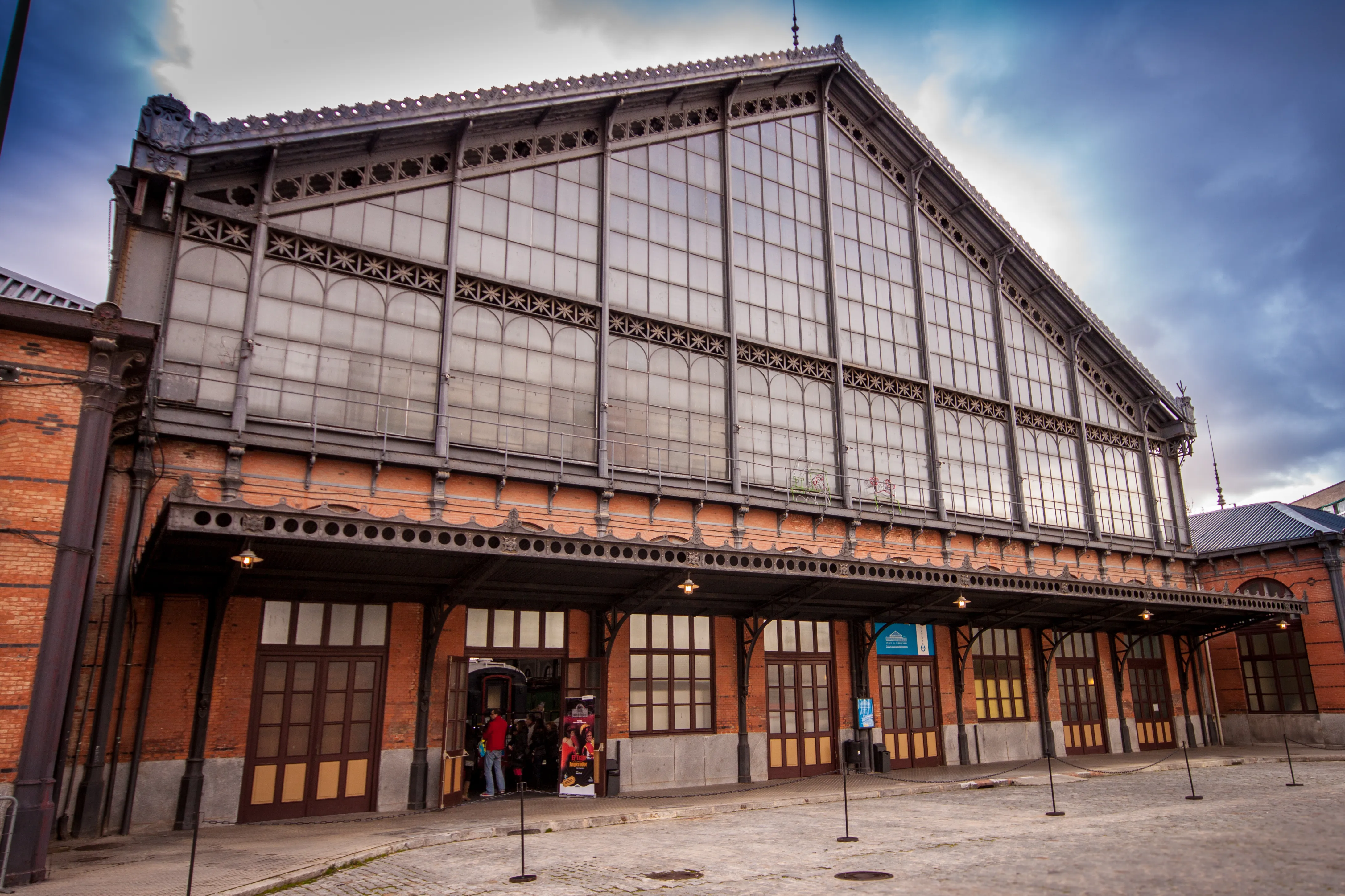 Large brick and glass building with a steeply pitched roof and arched windows, entrance doors with people entering