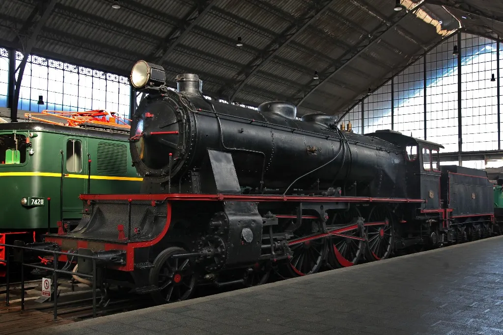 black steam locomotive with red trim on display inside a large hall with high arched ceiling and large windows
