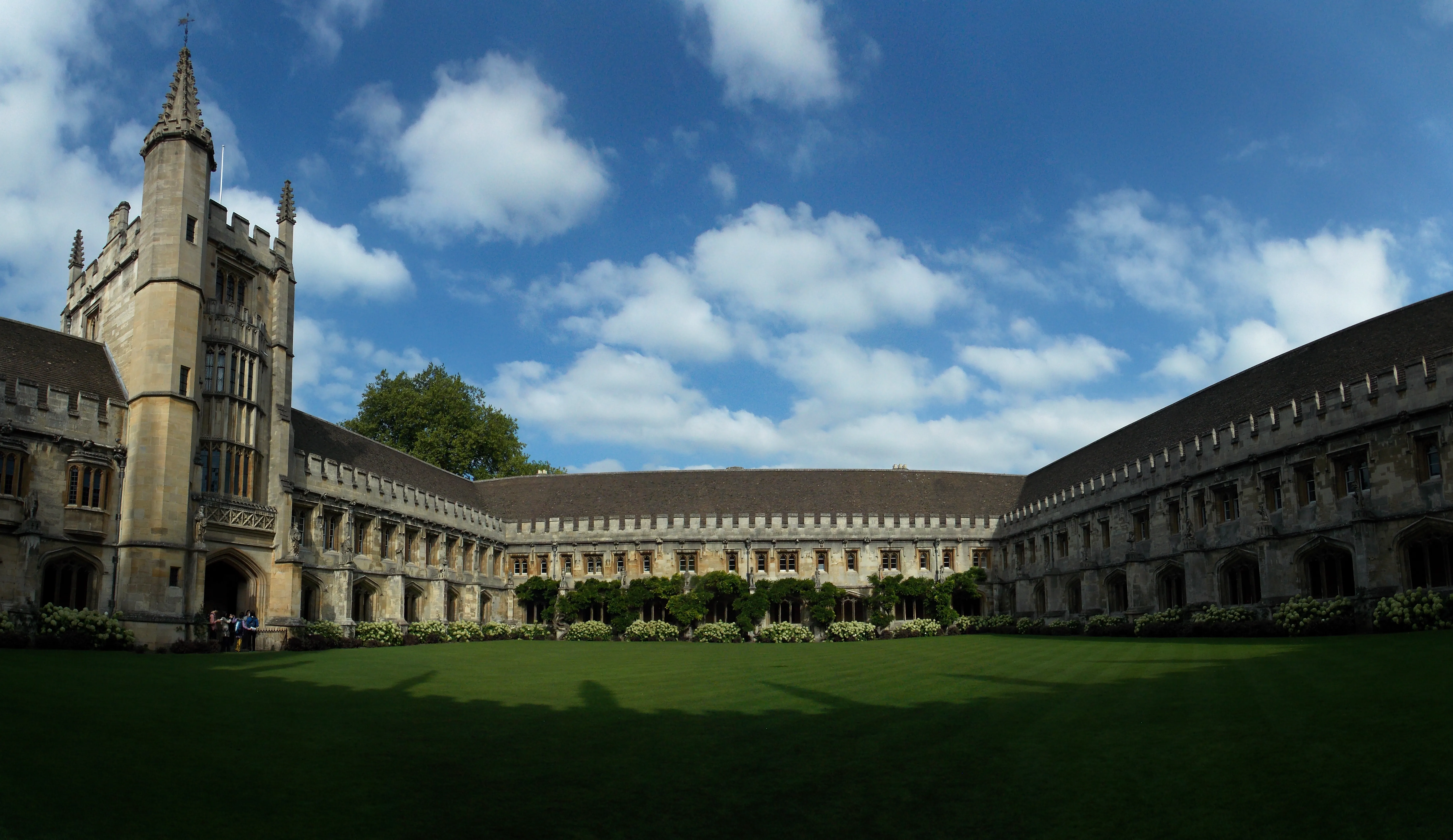 De Great Quadrangle van Magdalen College met de stenen toren met pinakels, kloostergangen en een groen gazon onder een blauwe lucht met witte wolken.