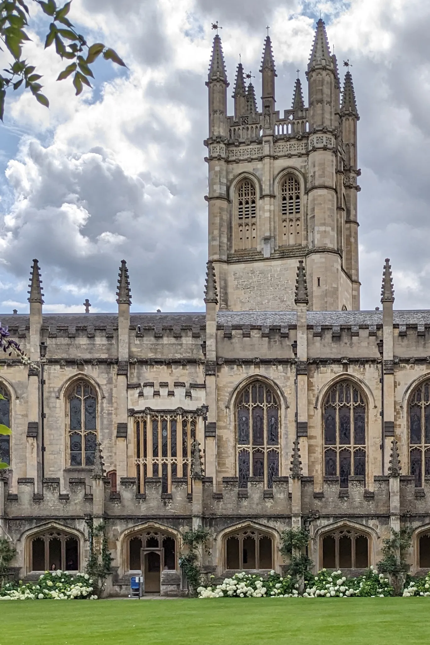 De Magdalen Tower die boven de gotische kloostergevel van Magdalen College in Oxford uitsteekt, met een groen gazon en witte bloemen op de voorgrond onder een bewolkte lucht.