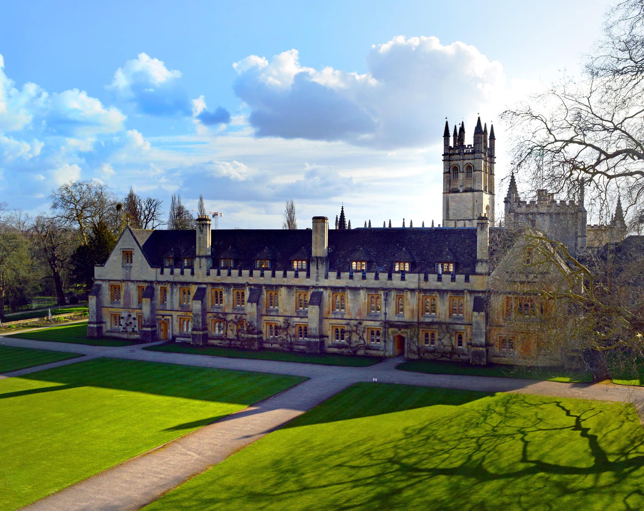 Stenen gebouwen van Magdalen College met de iconische Magdalen Tower die erboven uitsteekt, groene gazons en paden op de voorgrond onder een blauwe lucht met wolken.