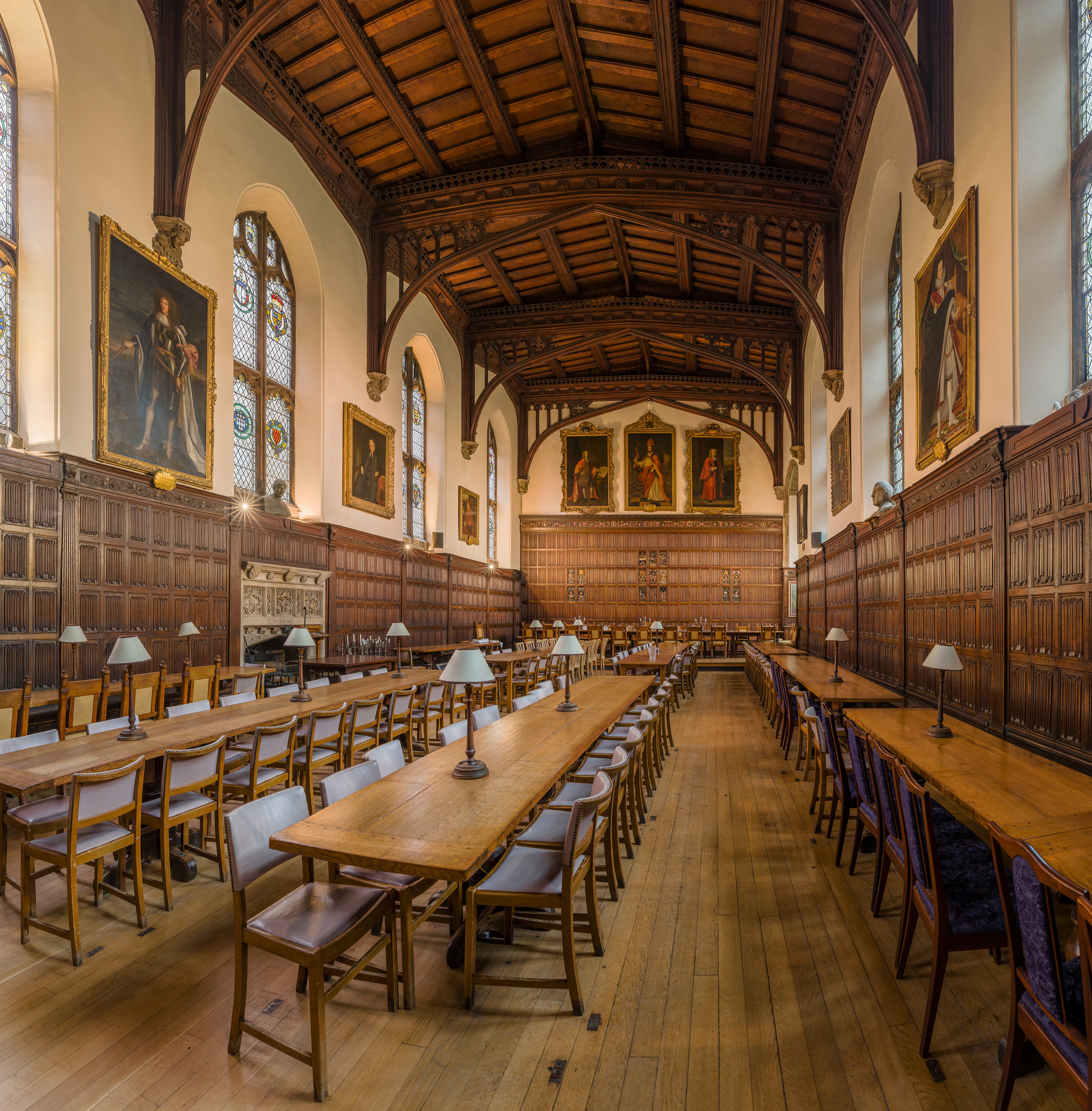 Een grandioze historische eetzaal met lange houten tafels en stoelen, een hoog plafond met zichtbare houten balken, houten lambrisering, grote portretschilderijen en glas-in-loodramen.
