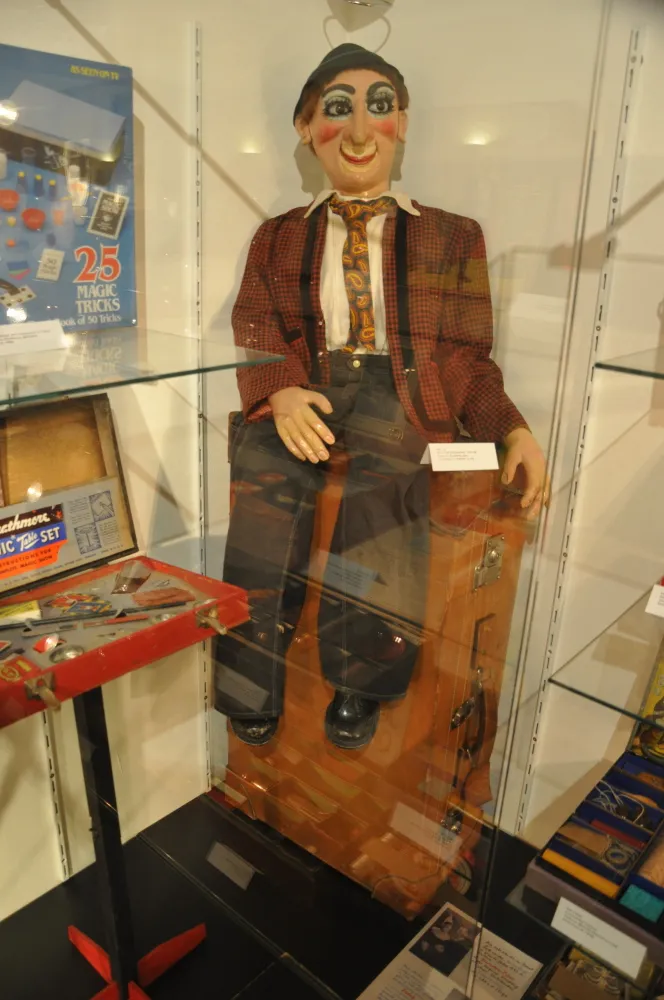 A mannequin dressed as a magician in a checkered jacket and bowler hat, sitting inside a wooden trunk on display in a glass case.